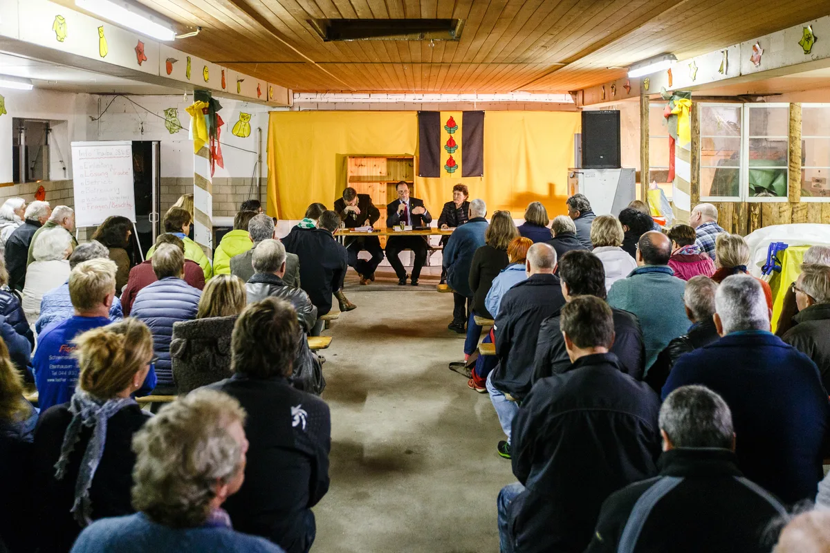 Ein Raum voll mit Leuten, die einem Mann am Mikrofon zuhören. 